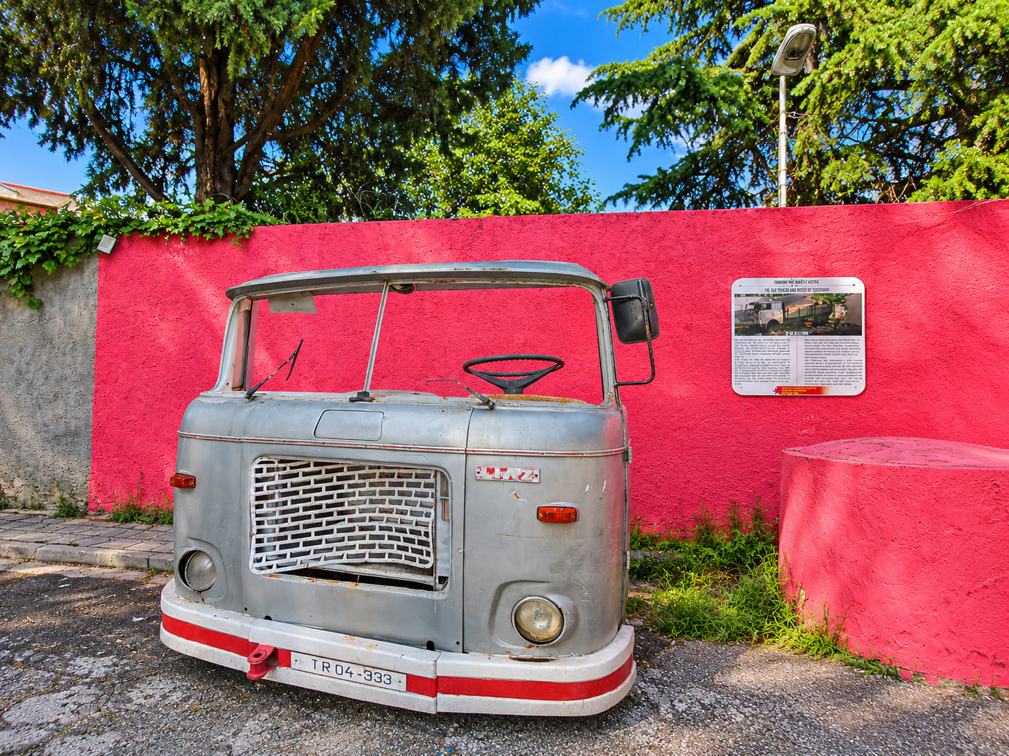 Vintage gray truck cab displayed in front of a bright pink wall at an outdoor historical exhibit in Tirana, Albania
