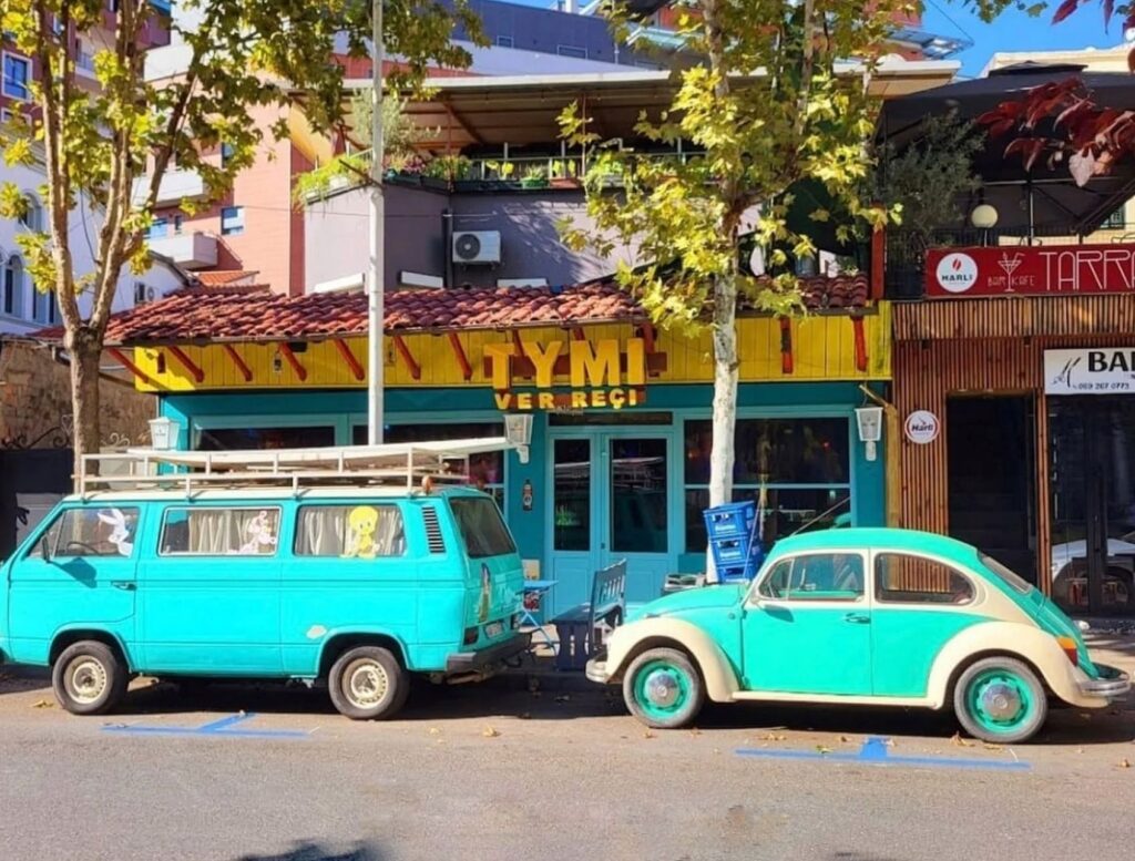 Yellow and teal storefront of Tymi restaurant in Tirana with a vintage van and VW Beetle parked outside