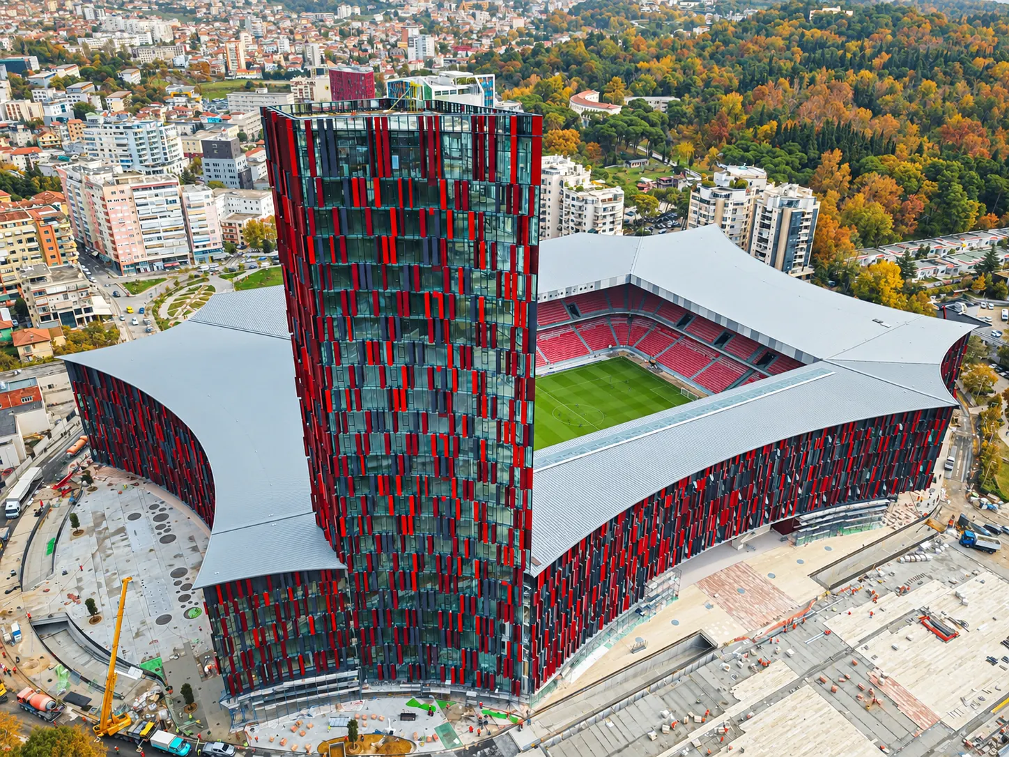 Aerial view of Air Albania Stadium in Tirana, featuring its red and black facade, football pitch, and surrounding cityscape with autumn trees
