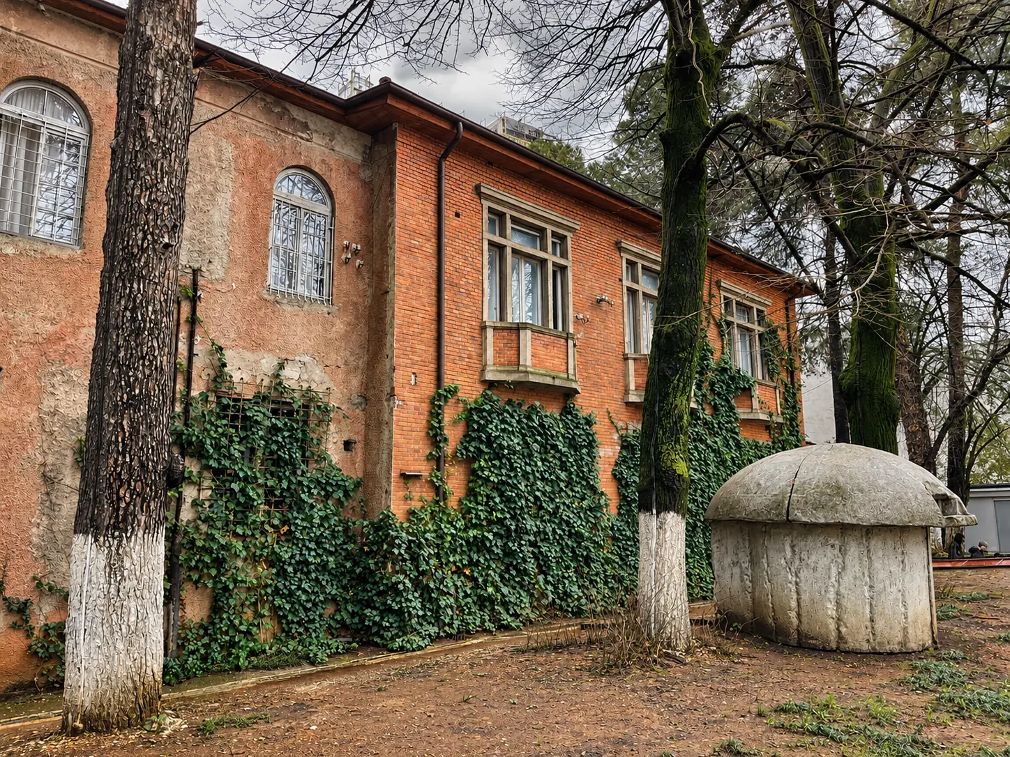 House of Leaves Museum Tirana, featuring the historic red-brick building covered in ivy, surrounded by trees, with a communist-era concrete bunker in the foreground